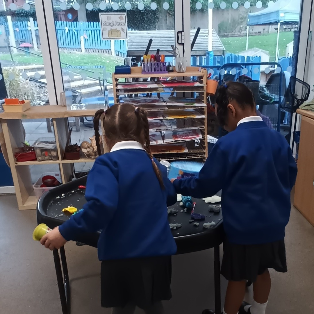 two children in blue school uniform playing at a tray with playdough on it. The photo is taken from behind