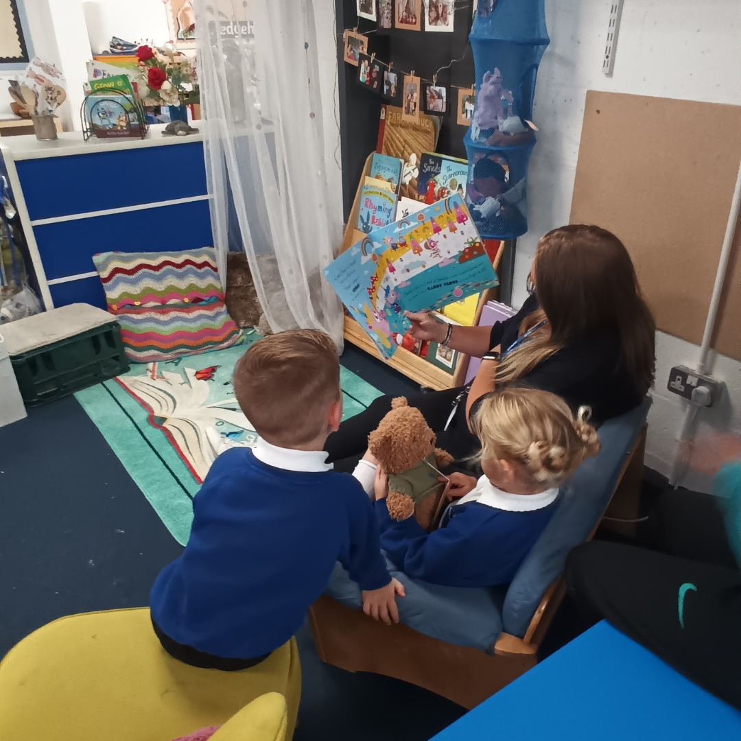 A boy and a girl in blue school uniform listening to a story being shared by an adult. One child is holding a teddy bear.